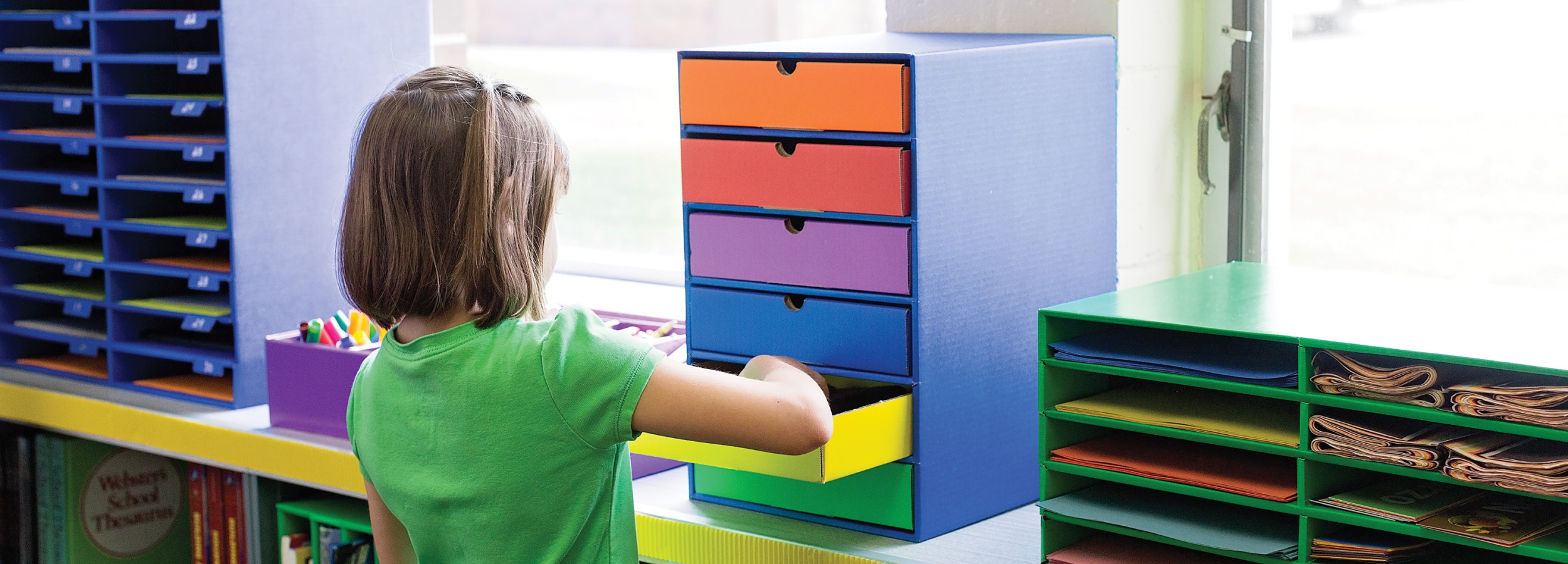 Child organizing colorful storage drawers in a bright classroom setting.